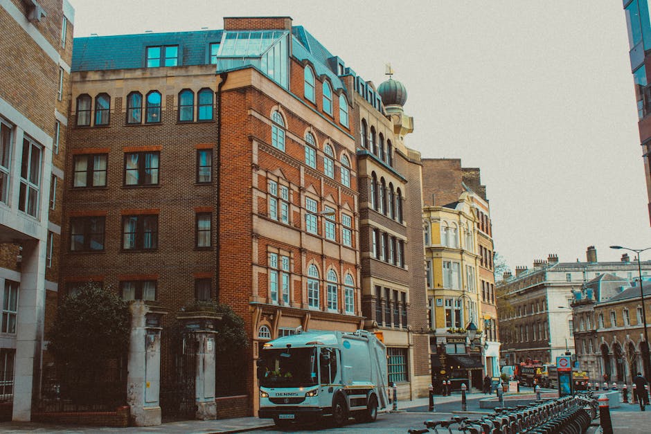 A residential street scene showing a white van parked outside a multi-storey building with large windows and decorative brickwork, which is part of a busy urban area. The van, likely used for furniture transport and home relocation, is positioned near the pavement, with its rear door open during the loading process. The loading area is adjacent to a narrow entrance, with packages and boxes possibly inside or being moved onto the vehicle. Surrounding the van are other buildings with mixed architectural styles, including brick and stucco facades, with additional vehicles and street furniture visible further along the road. The urban environment is well-lit with natural daylight, and the scene depicts the typical logistics involved in packing and moving services performed by companies such as Chiswick Man and Van, especially in areas with limited access requiring careful planning for house removals and furniture transport.
