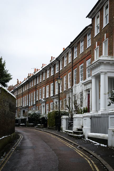 A narrow residential street in Chiswick with a row of traditional brick terraced houses featuring white window frames and decorative columns at the entrance, set against an overcast sky. The street is lined with pavement and has a slight curve, with a black lamppost positioned near the front of the properties. Some houses have small front gardens with bushes and snow-covered plants, and there is a low brick wall running along the pavement. The street appears to be in the early stages of a house removal, with a focus on urban residential relocation logistics. Chiswick Man and Van would assist with furniture transport, packing, and loading processes taking place in this setting, as part of house removals and home relocation services in tight access areas such as this.