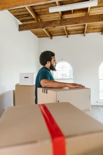 A man with dark curly hair and a beard, wearing a dark teal t-shirt, is standing inside a spacious, well-lit room with white walls and a wooden beamed ceiling. He is positioned behind several packed cardboard boxes, some of which are sealed with red tape. The boxes vary in size and are stacked on the floor, with one placed on a larger box. The man appears to be preparing for a house move or furniture transport, part of a home relocation process involving packing and loading. Behind him, there are two arched windows allowing natural light into the room, and the setting suggests interior prep work for professional removals, as seen in services by Chiswick Man and Van. The scene captures the logistical phase of packing and loading belongings, possibly for transportation in a van, which is typical during furniture transport or moving services related to house removals.