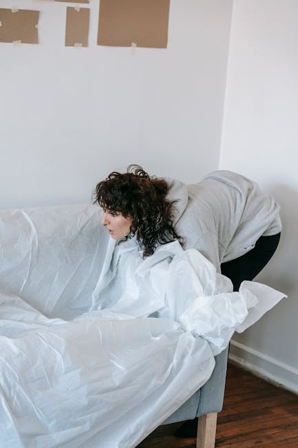 A woman with curly dark hair wearing a light grey hoodie is seen leaning over a white sofa in a room with white walls. She appears to be adjusting or preparing the sofa for moving, with white packing paper or protective wrapping covering parts of it. In the background, there are remnants of a previous wall or ceiling repair with brown paper or cardboard taped to the upper portion of the wall. The floor is dark wood, and the overall setting suggests a home environment during a house relocation or furniture packing process. Chiswick Man and Van, a professional removals company, may be involved in the furniture transport or packing activities related to house moving, as indicated by the context of the webpage on house removals and safe packing.