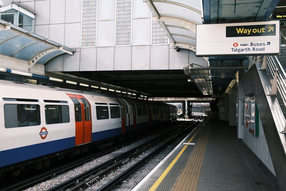 A London Underground train with a white, orange, and blue exterior is stationary on the underground platform beneath the station's curved, transparent roof. The platform features tactile paving along the edge and a sign indicating the way out towards Talgart Road and buses overhead. Inside the station, there is a staircase with metal railings leading up to street level. The surroundings include concrete walls and structural supports, with lighting fixtures attached to the ceiling. The scene captures an urban transport environment potentially involved in a home relocation process, with the station sign and platform layout reflecting a typical setting where moving services such as furniture transport and packing can be coordinated by companies like Chiswick Man and Van during a moving day.