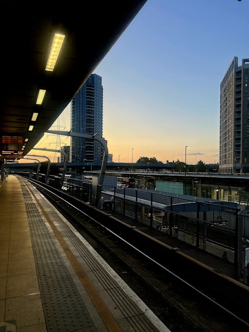 Exterior view of Chiswick Park station platform during early evening with the sun setting in the background. The platform is partially covered with a black overhead canopy featuring built-in linear lighting. To the left, the train tracks run parallel to the platform, which is lined with tactile paving and safety railings. In the distance, modern high-rise buildings and the skyline of London are visible against a clear sky, indicating an urban environment suitable for home relocation and furniture transport services. The scene captures the calm atmosphere of a busy transportation hub, highlighting the importance of efficient moving logistics for house removals near Chiswick. Chiswick Man and Van regularly assists with packing and loading during here, ensuring smooth transitions for clients during their house moves.