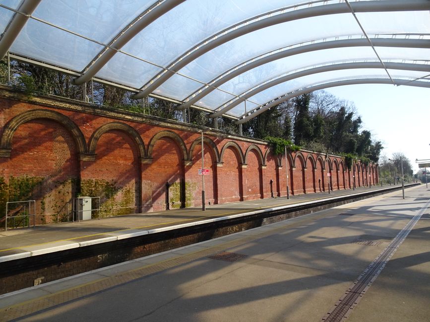 A London Underground train with a white, orange, and blue exterior is stationary on the underground platform beneath the station's curved, transparent roof. The platform features tactile paving along the edge and a sign indicating the way out towards Talgart Road and buses overhead. Inside the station, there is a staircase with metal railings leading up to street level. The surroundings include concrete walls and structural supports, with lighting fixtures attached to the ceiling. The scene captures an urban transport environment potentially involved in a home relocation process, with the station sign and platform layout reflecting a typical setting where moving services such as furniture transport and packing can be coordinated by companies like Chiswick Man and Van during a moving day.