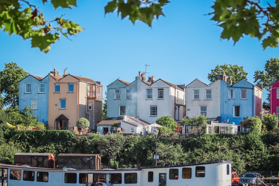 A row of four multi-storey houses painted in pastel shades of blue, beige, white, and pink, situated on an elevated area with lush green trees and shrubs in front. The houses feature various window st