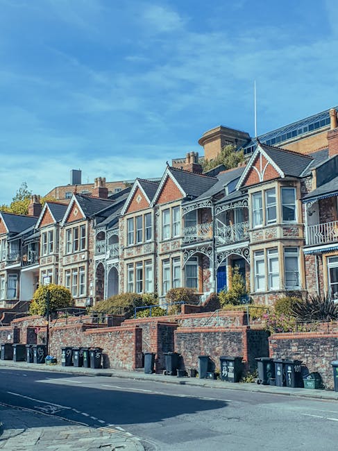 A row of Victorian terraced houses in Chiswick with ornate ironwork balconies, large bay windows, and brick facades, situated on a sloped street with a brick retaining wall and garden beds in front. The pavement is lined with multiple black wheelie bins, and a clear blue sky is visible overhead. This residential scene illustrates typical housing that might require professional house removals or home relocation services, such as those provided by Chiswick Man and Van, to facilitate packing, furniture transport, and other moving logistics.