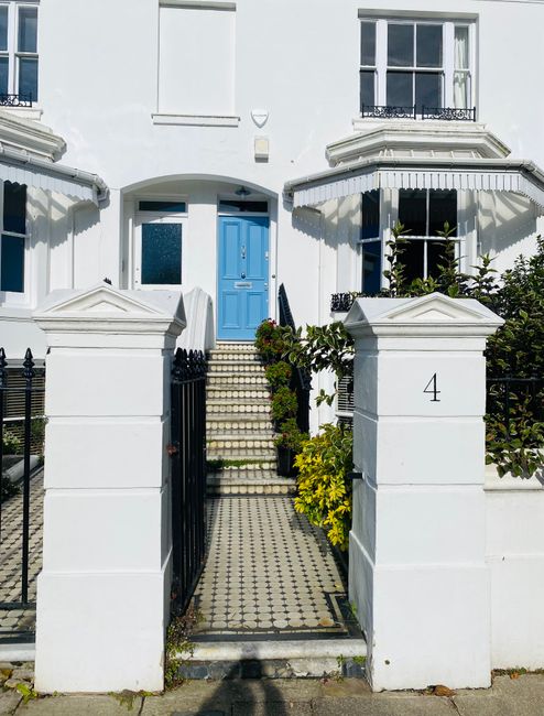 A front view of a white Victorian-style house in Chiswick with a bright blue front door accessed by a narrow tiled pathway. The pathway is flanked by white stone pillars with black metal gates, leading up to a staircase with small square tiles, passing through a small garden area with green plants and yellow flowers. The house features large sash windows with black wrought-iron decorative balcony railings, and two white bay windows with striped fabric awnings on either side of the door. The exterior is well-maintained, with clean white walls and detailed architectural features. This image captures a typical residential property suitable for house removals or home relocation services, as provided by Chiswick Man and Van, and illustrates the entry process for furniture transport or packing and moving activities at this property.