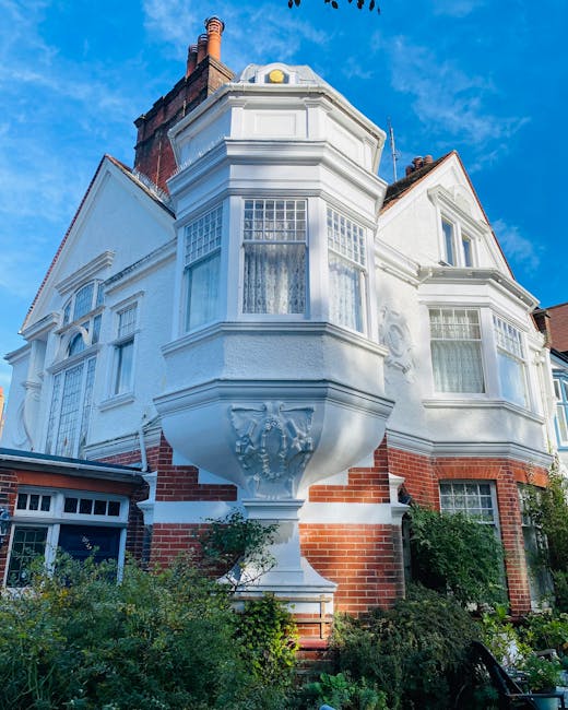 A row of Victorian terraced houses in Chiswick with ornate ironwork balconies, large bay windows, and brick facades, situated on a sloped street with a brick retaining wall and garden beds in front. The pavement is lined with multiple black wheelie bins, and a clear blue sky is visible overhead. This residential scene illustrates typical housing that might require professional house removals or home relocation services, such as those provided by Chiswick Man and Van, to facilitate packing, furniture transport, and other moving logistics.